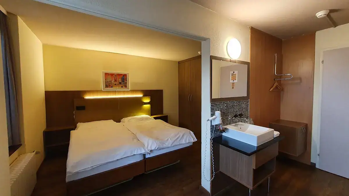 Hotelzimmer mit Doppelbett, Nachttischen, Lampe, Waschbecken und Kleiderschrank.